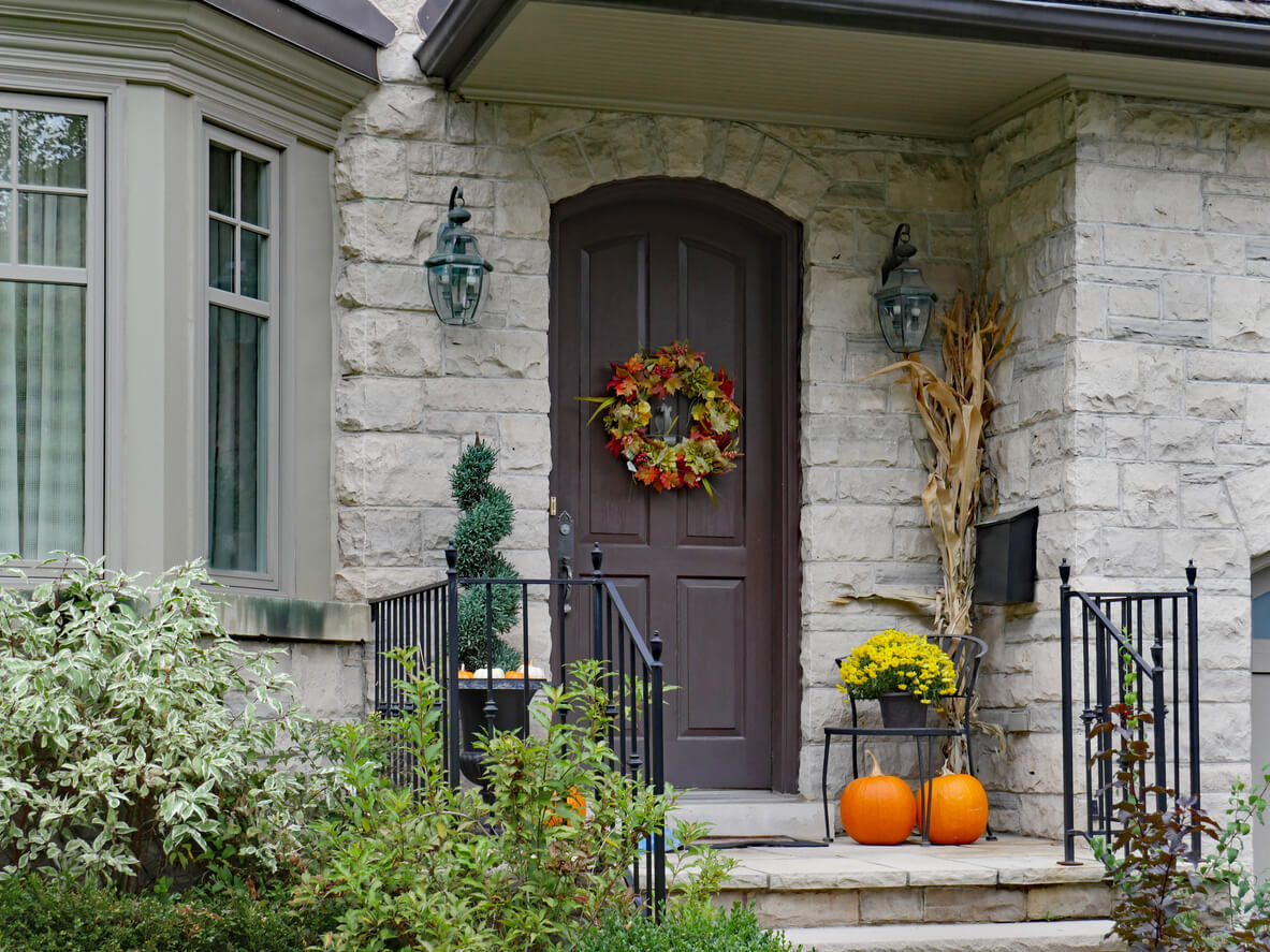 A fall-colored wreath on a brown door of a stoned house. There are pumpkins, and flowers next to the door by the stoop.