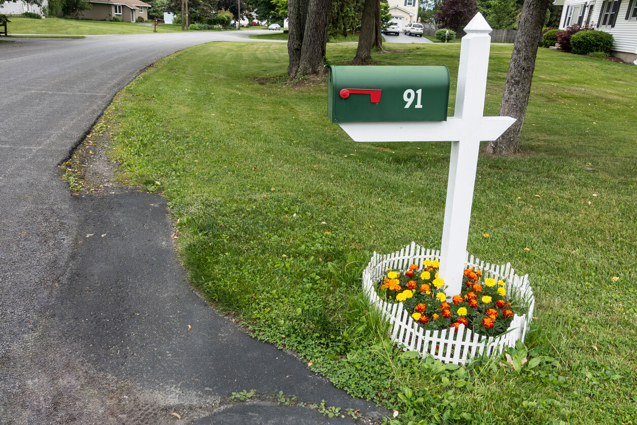 Green mailbox at the end of a driveway with a small garden of yellow and orange flowers surrounded by a small picket white fence at the bottom of the post