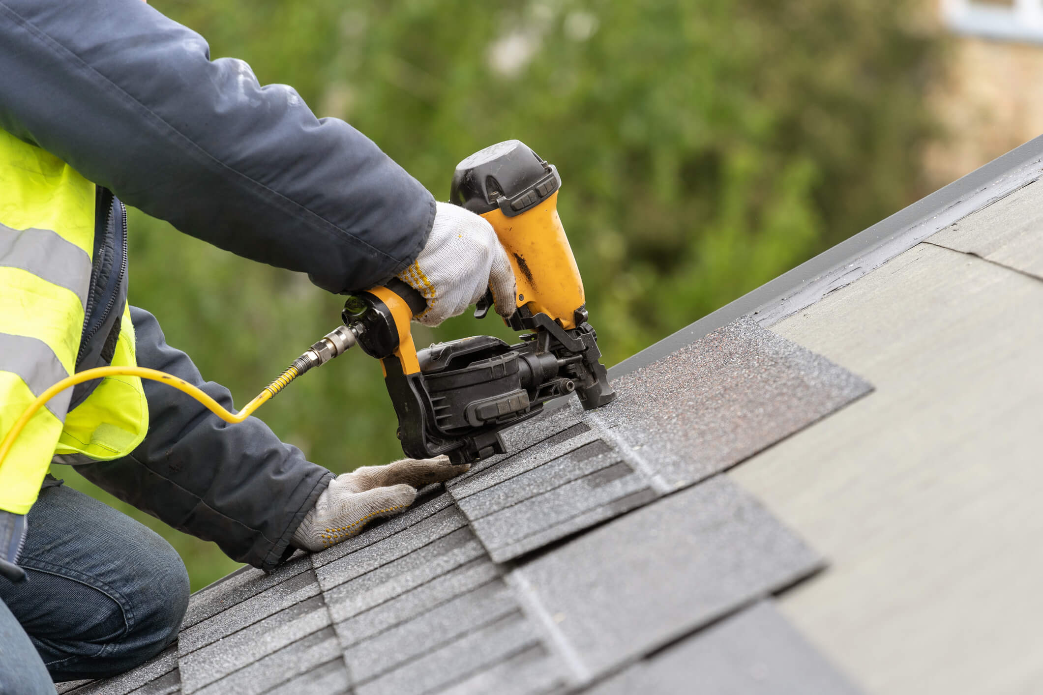 person using nail gun to install asphalt shingles on roof